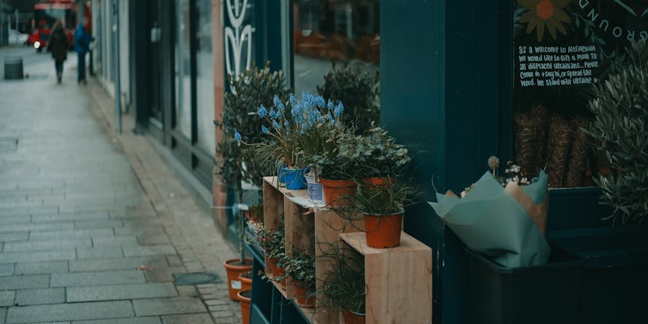 A display of potted plants and floral arrangements outside a flower shop, arranged on light wooden shelves and in a large dark container; the plants include blue forget-me-nots, green foliage, and sma