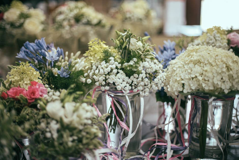 A collection of floral arrangements featuring white hydrangeas, pink roses, blue irises, and delicate white baby's breath, displayed in clear glass vases wrapped with pastel pink and white ribbons. Th