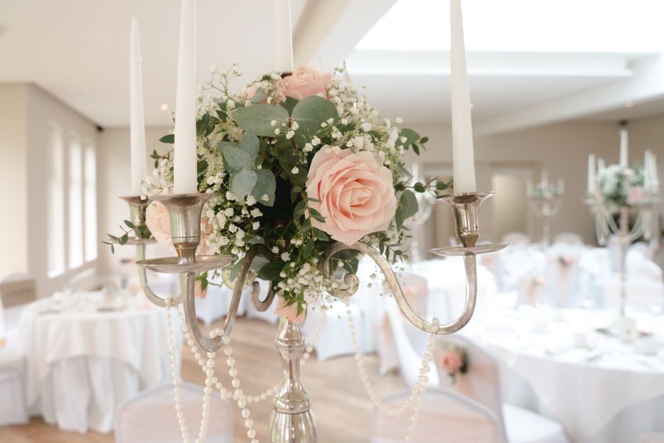 A floral arrangement featuring light pink roses, white baby's breath, and eucalyptus leaves, arranged in a round bouquet style. The flowers are displayed on a silver candelabrum decorated with hanging