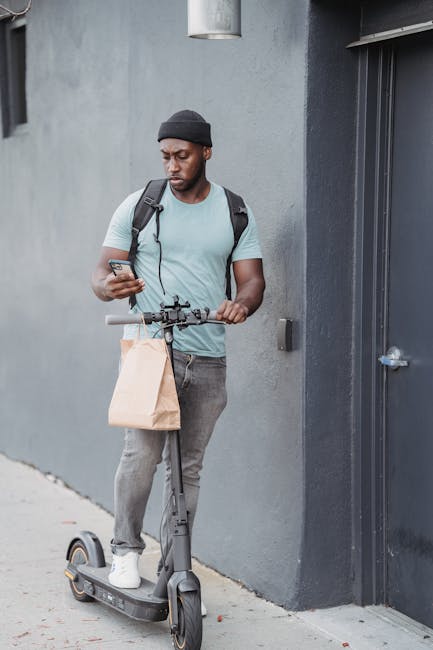 A young man with a dark beanie and light blue t-shirt stands outdoors next to a gray wall, holding a smartphone in one hand and a small paper bag hanging from the handlebar of an electric scooter he i