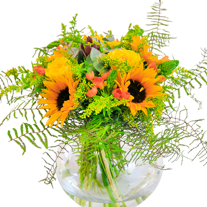 Upon first glance, this elaborate Watford bouquet projects sunlit positivity as it rests within its rounded glass vase. Majestic sunflowers dominate, their radiant petals and deep, central disks capturing the attention and suggesting endless warmth, much like the inviting parks and open spaces scattered throughout the town. Flanking these are clusters of softly curled yellow garden roses, their petals closely bound in romantic spirals and perfuming the air, echoing the gentle charm of Oxhey Village in the early summer months. Scarlet-red hypericum berries, dotted throughout, gleam with a jewel-like quality, introducing playful contrast to the yellow spectrum. Lush feathery ferns create a sense of movement, their soft lines reminiscent of wild greenery trailing alongside Watford's leafy footpaths. Light green filler, possibly goldenrod dusted with numerous tiny flowers, and cool eucalyptus leaves thread through the display, adding a lively sense of depth. The stems, visible through the clear water of the vase, showcase the arrangement's natural architecture and freshness. Gentle sunlight caresses the petals and leaves, casting mellow reflections and animating the bouquet with a sense of cheer. This is a creation well suited for commemorating milestones, expressing gratitude, or delivering joy to a loved one in WD17-an elegant statement of nature's bounty and local pride, expertly fashioned by Watford Florist.